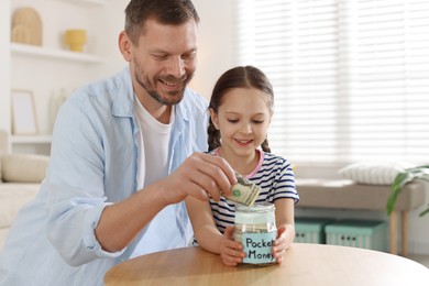 Photo of Father and daughter putting pocket money into jar at table indoors