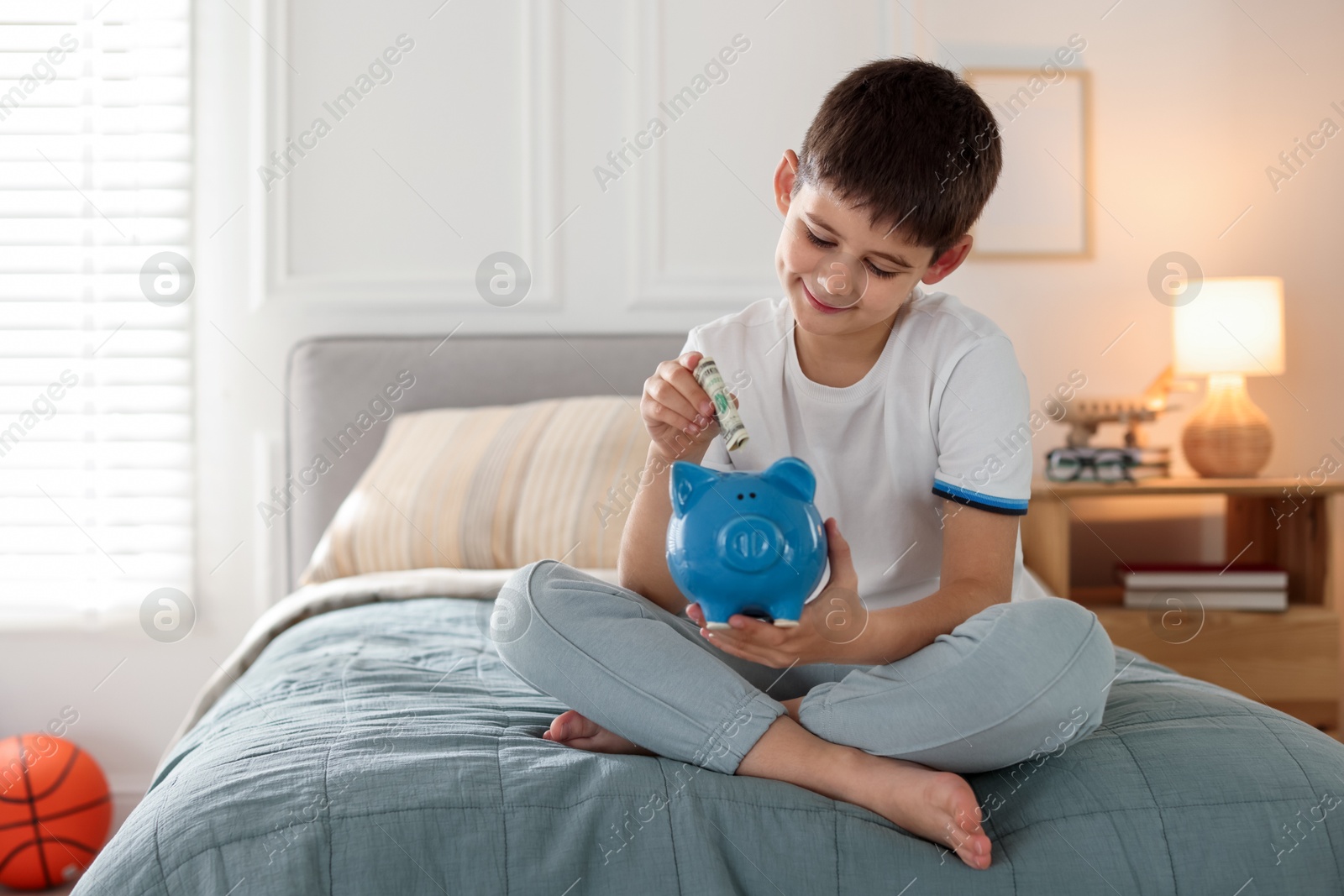 Boy putting pocket money into piggybank on bed at home. Space for text Photo of Boy putting pocket money into piggybank on bed at home. Space for text