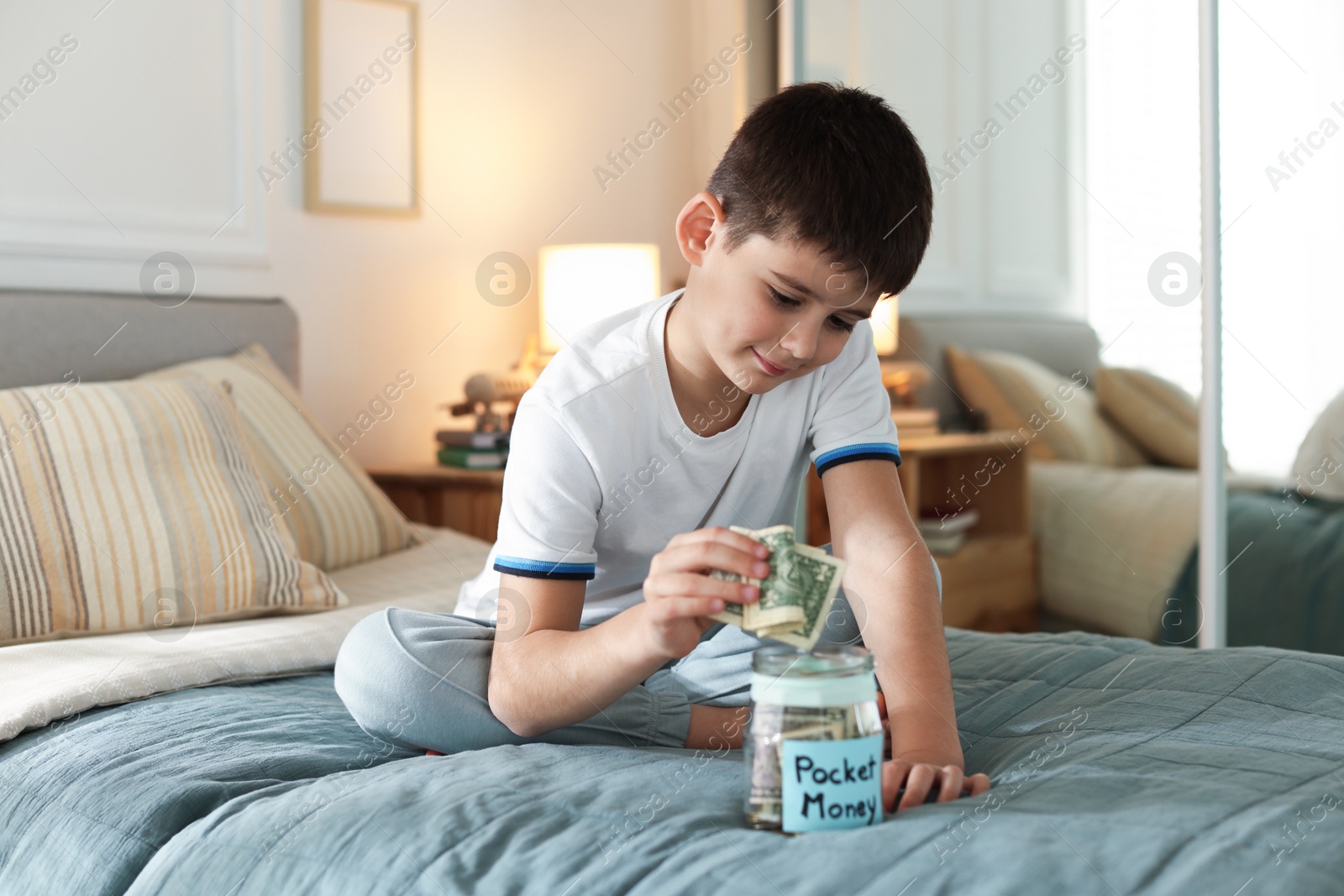 Boy putting pocket money into jar on bed at home Photo of Boy putting pocket money into jar on bed at home
