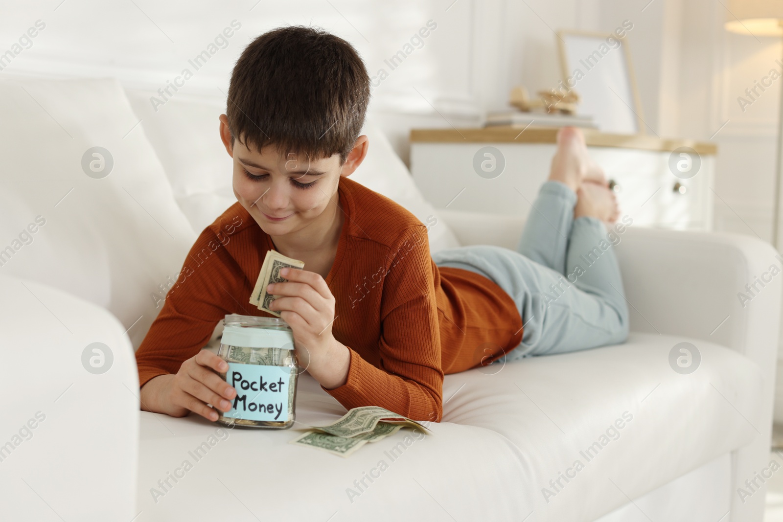 Boy putting his pocket money into jar on sofa at home Photo of Boy putting his pocket money into jar on sofa at home