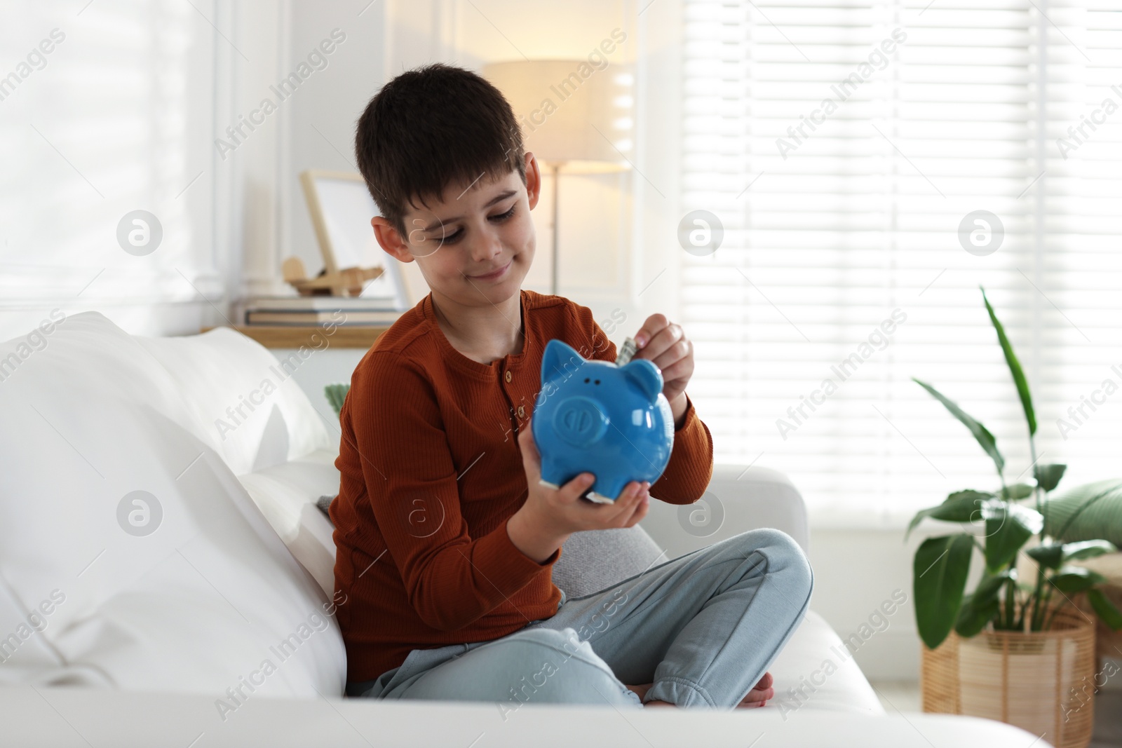 Boy putting his pocket money into piggybank on sofa at home Photo of Boy putting his pocket money into piggybank on sofa at home
