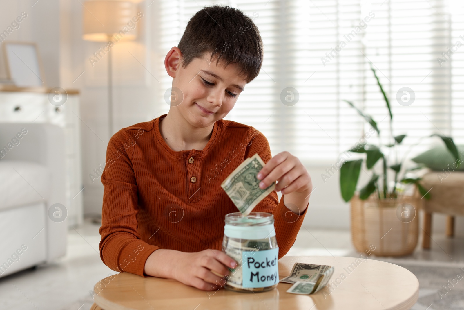 Boy putting his pocket money into jar at table indoors Photo of Boy putting his pocket money into jar at table indoors