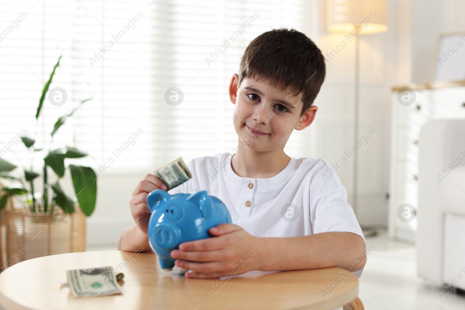 Boy putting pocket money into piggybank at table indoors Photo of Boy putting pocket money into piggybank at table indoors