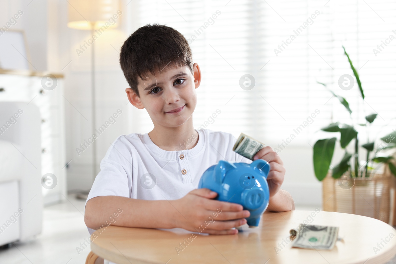 Boy putting pocket money into piggybank at table indoors Photo of Boy putting pocket money into piggybank at table indoors