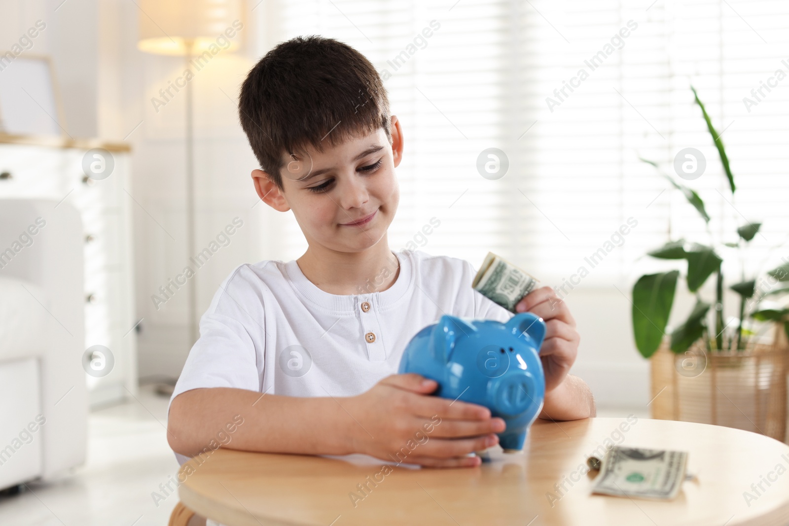 Boy putting pocket money into piggybank at table indoors Photo of Boy putting pocket money into piggybank at table indoors