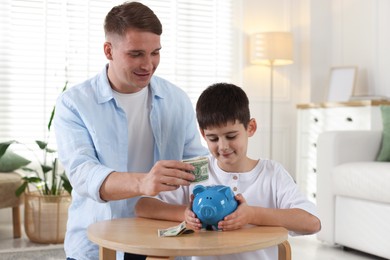 Photo of Father and son putting pocket money into piggybank at table indoors