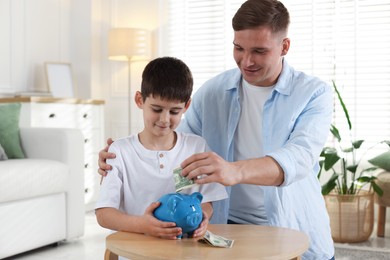Photo of Father and son putting pocket money into piggybank at table indoors