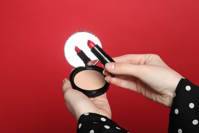 Photo of Woman holding open face powder with mirror and lipstick on red background, closeup