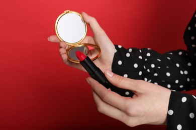 Photo of Woman holding open pocket mirror and lipstick on red background, closeup