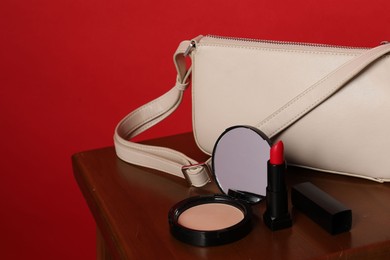 Photo of Open face powder with mirror, lipstick and bag on wooden stool against red background, closeup