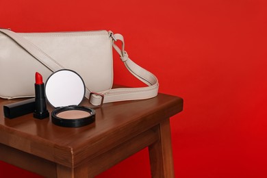 Photo of Open face powder with mirror, lipstick and bag on wooden stool against red background, space for text