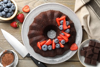 Photo of Tasty chocolate bundt cake with berries and knife on wooden table, flat lay