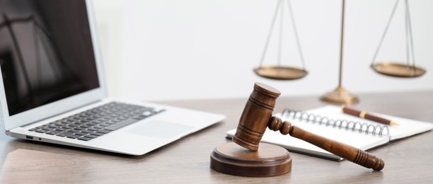 Photo of Laptop, judge's gavel and scales on wooden table in office, closeup