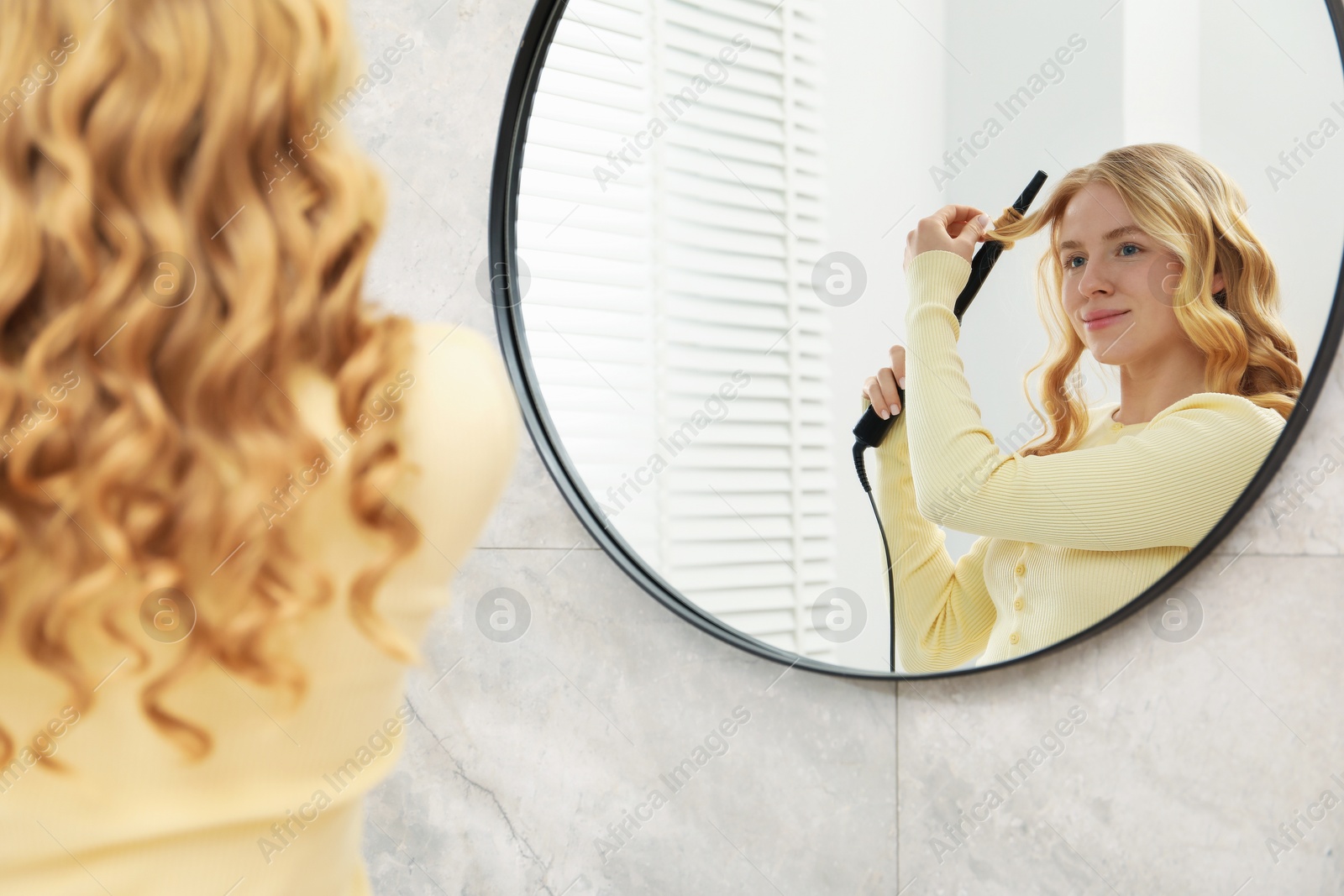 Woman curling her hair near mirror indoors Photo of Woman curling her hair near mirror indoors