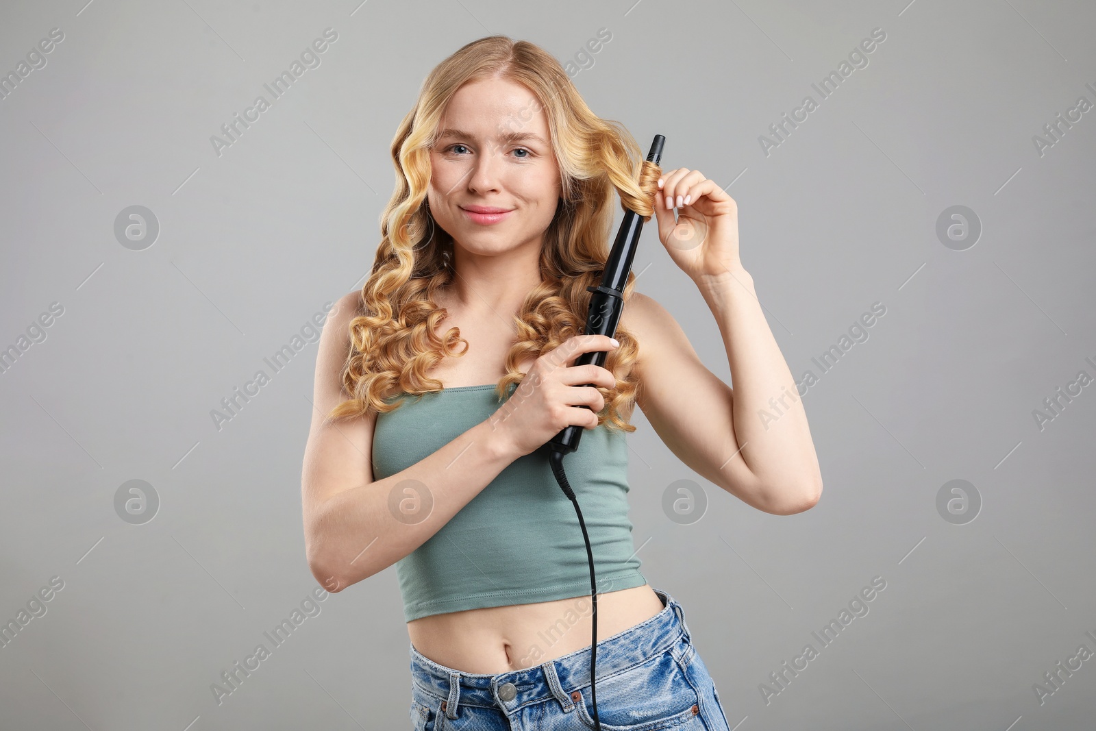 Woman curling her hair on grey background Photo of Woman curling her hair on grey background