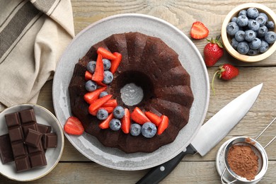 Photo of Tasty chocolate bundt cake with berries and knife on wooden table, flat lay