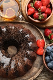 Photo of Tasty chocolate bundt cake with glaze, tea and berries on wooden table, flat lay
