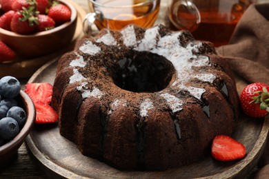 Photo of Tasty chocolate bundt cake with glaze, tea and berries on wooden table, closeup