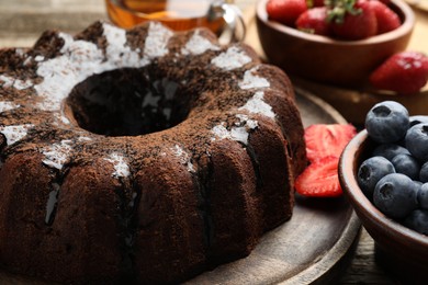 Photo of Tasty chocolate bundt cake with glaze, tea and berries on wooden table, closeup