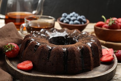 Photo of Tasty chocolate bundt cake with glaze, tea and berries on wooden table, closeup