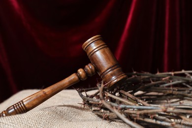 Photo of Judge's gavel and crown of thorns on burlap fabric against red curtain, closeup