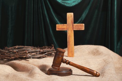 Photo of Judge's gavel, wooden cross and crown of thorns on burlap fabric against teal curtain