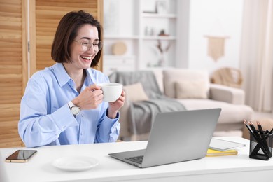 Photo of Smiling woman with cup of hot drink having videochat by laptop at desk in home office