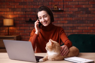 Photo of Smiling woman talking by smartphone with laptop and cat at desk in home office