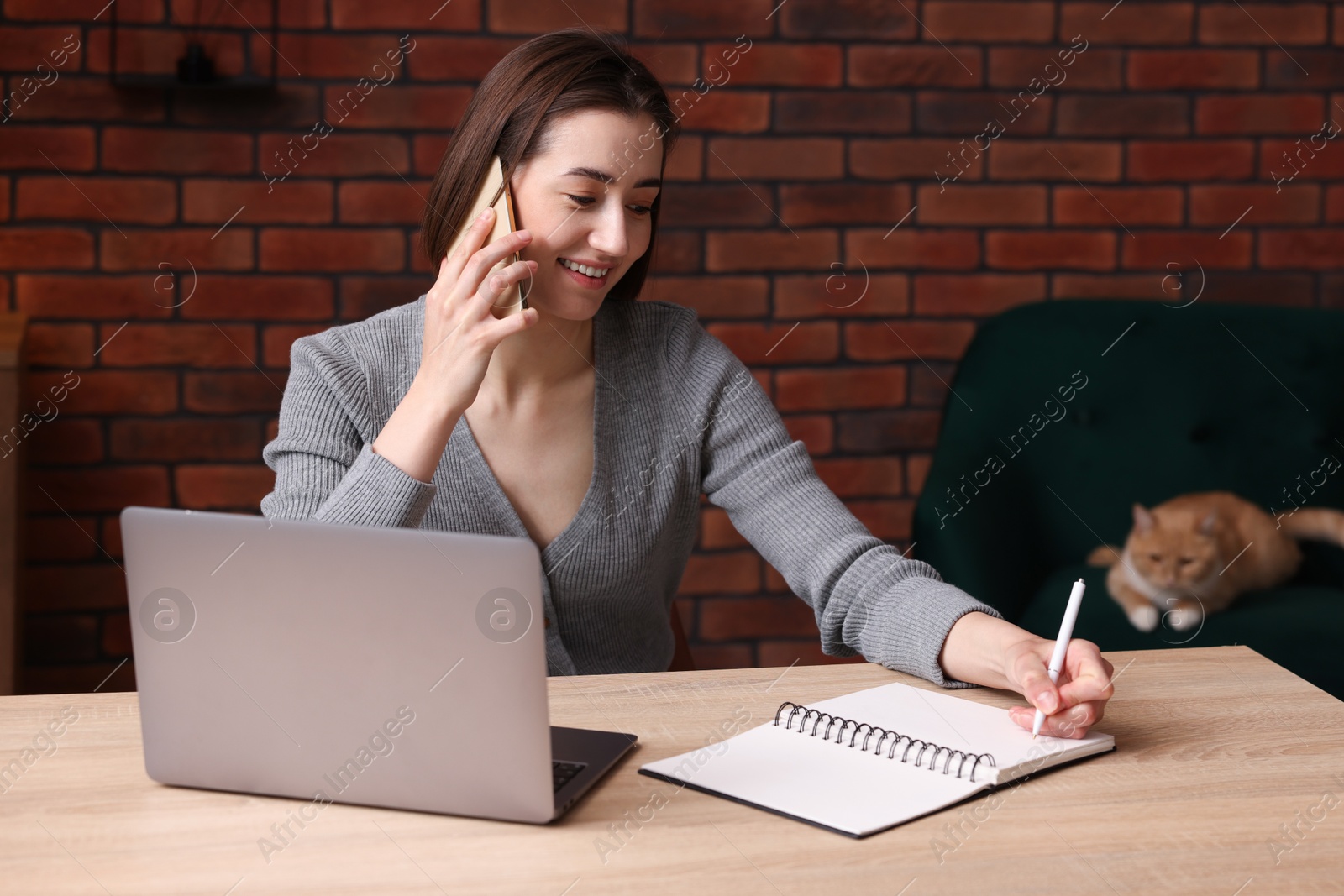 Smiling woman talking by smartphone at desk with laptop in home office Photo of Smiling woman talking by smartphone at desk with laptop in home office