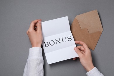 Image of Woman holding paper with word Bonus at grey table, top view