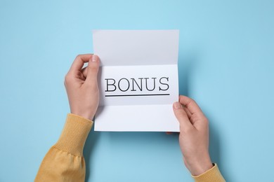 Image of Woman holding paper with word Bonus at light blue table, top view