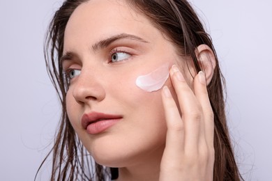 Photo of Beautiful woman applying skincare product onto her face on light grey background, closeup