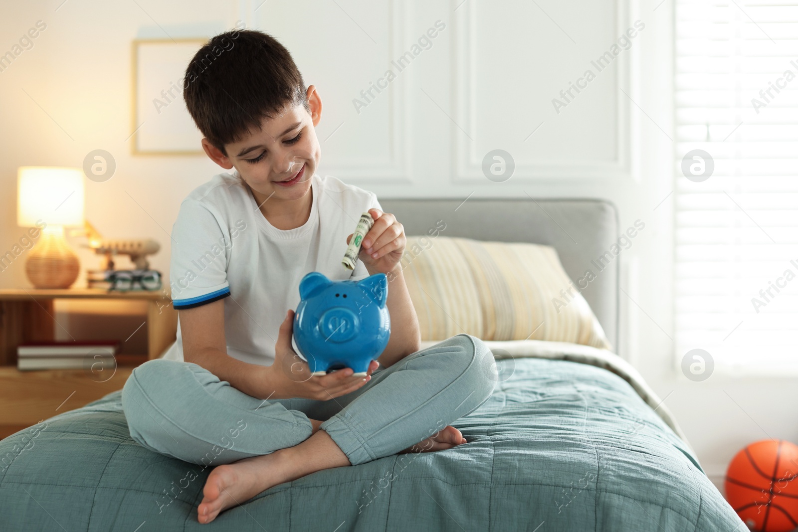 Boy putting pocket money into piggybank on bed at home. Space for text Photo of Boy putting pocket money into piggybank on bed at home. Space for text