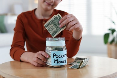 Photo of Boy putting his pocket money into jar at table indoors, closeup