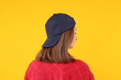 Photo of Young woman wearing stylish black baseball hat on yellow background, back view