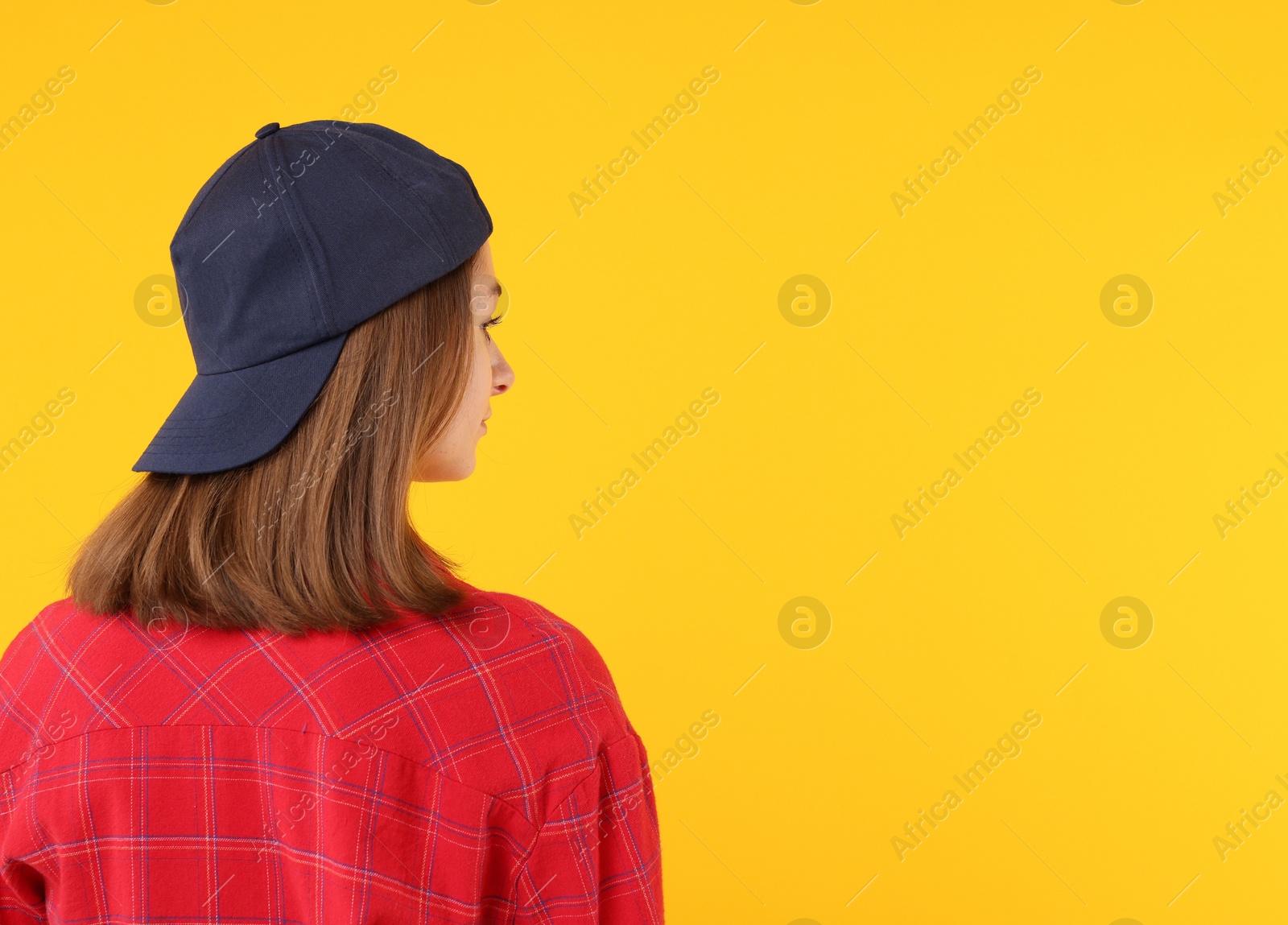 Photo of Young woman wearing stylish black baseball hat on yellow background, back view. Space for text