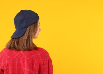 Photo of Young woman wearing stylish black baseball hat on yellow background, back view. Space for text
