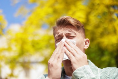 Photo of Young man with paper tissue suffering from seasonal pollen allergy near tree on spring day. Space for text