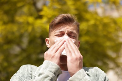 Photo of Young man with paper tissue suffering from seasonal pollen allergy near tree on spring day