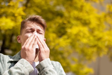 Photo of Young man with paper tissue suffering from seasonal pollen allergy near tree on spring day. Space for text