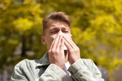 Photo of Young man with paper tissue suffering from seasonal pollen allergy near tree on spring day
