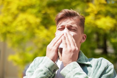 Photo of Young man with paper tissue suffering from seasonal pollen allergy near tree on spring day. Space for text