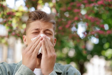 Photo of Young man with paper tissue suffering from seasonal pollen allergy near blossoming tree on spring day. Space for text