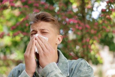 Photo of Young man with paper tissue suffering from seasonal pollen allergy near blossoming tree on spring day