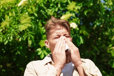 Photo of Young man with paper tissue suffering from seasonal pollen allergy near blossoming tree on spring day