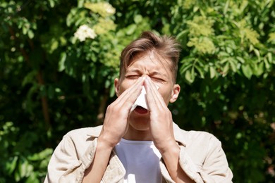 Photo of Young man with paper tissue suffering from seasonal pollen allergy near blossoming tree on spring day