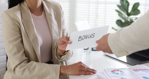 Photo of Employee receiving envelope with word Bonus from boss in office, closeup