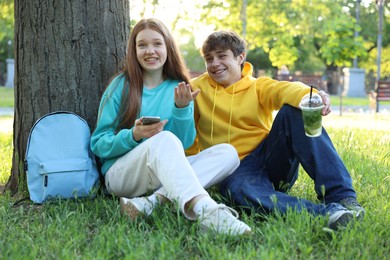 Photo of Teenage girl and boy with smartphone and drink on green grass in park