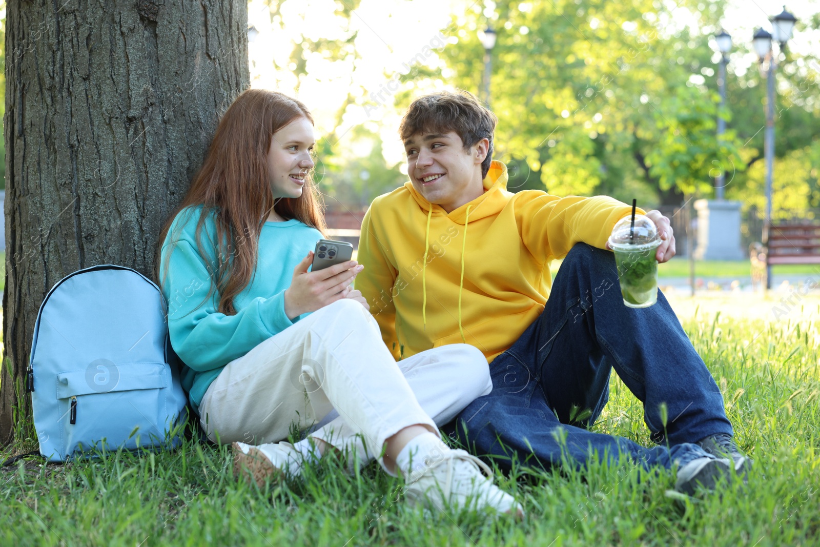 Photo of Teenage girl and boy with smartphone and drink on green grass in park