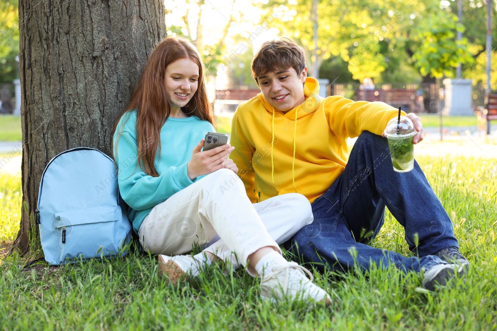 Teenage girl and boy with smartphone and drink on green grass in park Photo of Teenage girl and boy with smartphone and drink on green grass in park